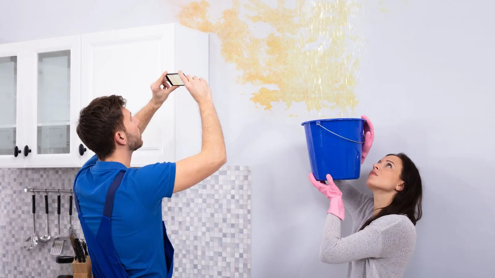 Homeowner photographing a water-stained wall while a bucket catches drips
